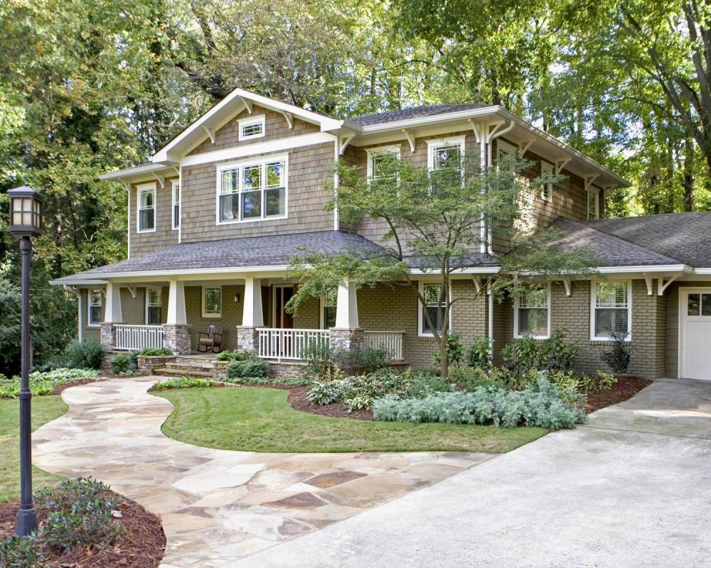 Two-story craftsman home with shingle siding, wide front porch, stone accents, and a curved flagstone walkway surrounded by mature trees — example of residential renovation done right.